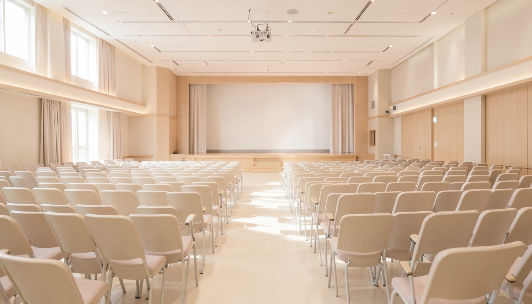 柔らかな光に照らされた広い学会会場の風景。淡いベージュと白を基調とした空間に、整然と並ぶ椅子とステージが穏やかな雰囲気を醸し出している。